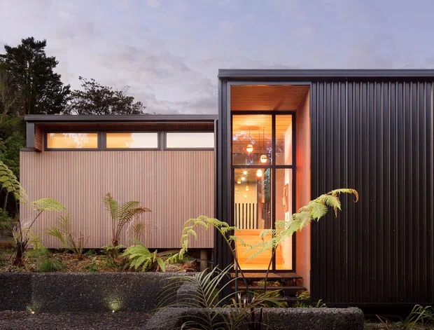 Exterior view of a compact modern building composed of dark corrugated metal cladding and a lighter vertically ribbed section. Warm light glows through narrow clerestory windows and a tall glazed entrance, with planted ferns and a dark retaining wall in the foreground.