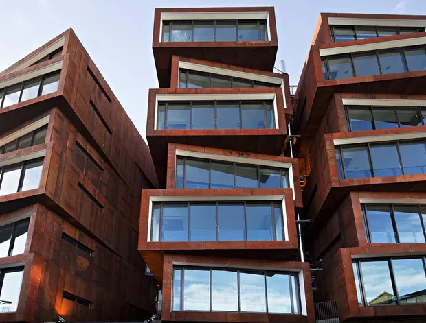 Exterior architectural photograph of a multi-storey contemporary building composed of several staggered, projecting volumes. The facade is clad in dark reddish-brown timber or weathered wood panels, with extensive floor-to-ceiling glazing, deep rectangular window surrounds, and stacked terraces or balconies. The upward-looking view emphasizes the layered massing and modern urban form.