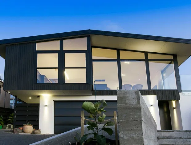 Exterior view of a contemporary two-storey residence at dusk, featuring dark vertical cladding, broad floor-to-ceiling glazing, a low-pitched projecting roof, and a ground-level garage. A concrete privacy wall and landscaped planting occupy the foreground.