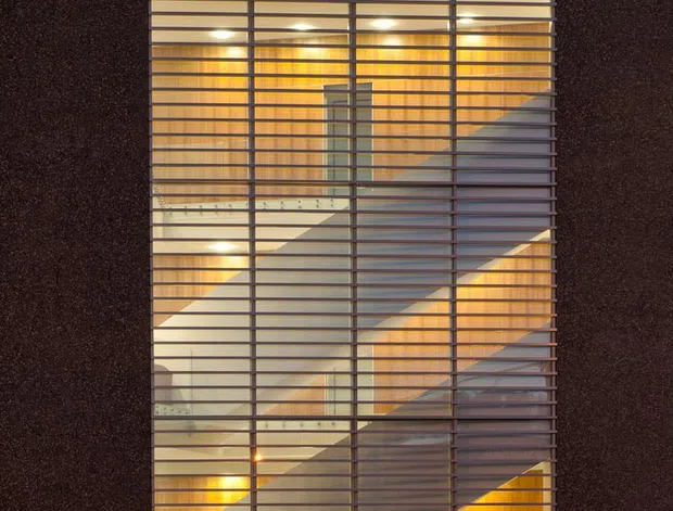 A dusk exterior view of a modern multi-storey building facade. A tall, narrow glazed stairwell or circulation core is set into dark textured cladding, screened by closely spaced horizontal metal louvres. Warm interior lighting reveals a large zigzagging stair and pale internal surfaces behind the glazing. The surrounding facade is largely dark and minimalist, with a paved parking area in the foreground.