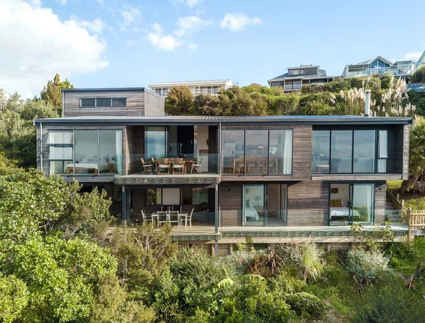 A contemporary multi-level residential house set into a lush, sloping coastal landscape. The home has a long, low horizontal form with dark timber-look cladding, extensive floor-to-ceiling glazing, projecting balconies and terraces, and a flat or very low-pitched roof. Native planting surrounds the building, with other hillside homes visible behind.