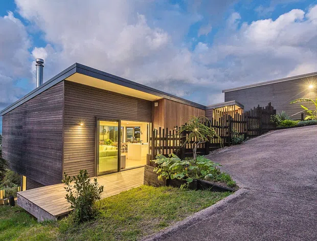 Exterior view of a contemporary single-storey home on a sloped site, featuring dark horizontal cladding, a low-pitched roof, warm recessed soffit lighting, large glazed doors, a timber deck, and landscaped planting. A separate matching volume and screened fencing are visible uphill beside the driveway.