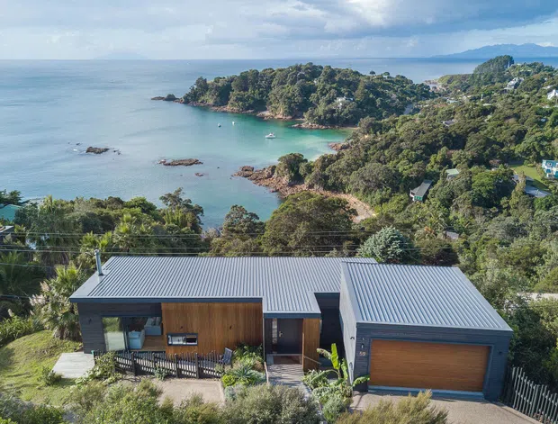 Aerial view of a contemporary single-storey coastal home with multiple low-pitched corrugated metal roof volumes, dark exterior cladding, warm timber garage and entry doors, and large glazing. The house sits on a landscaped sloping site overlooking a sheltered bay, rocky shoreline, dense vegetation, and distant hills.