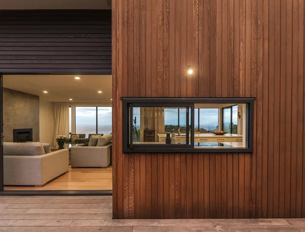 Exterior view of a contemporary residence clad in vertical dark timber, with a recessed glazed opening and a large sliding glass door revealing a warm living room inside. The interior includes light upholstered seating, timber flooring, a fireplace, recessed ceiling lights, and broad views toward a coastal landscape. A timber deck spans the foreground.