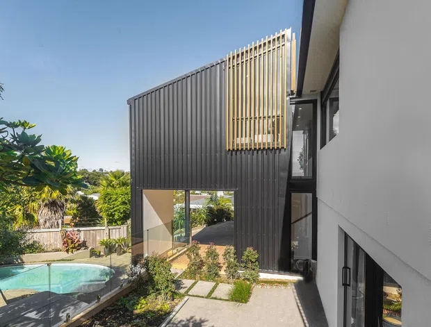 Exterior architectural view of a contemporary multi-level home, showing dark vertical cladding, a projecting upper-level screen of slim timber-coloured battens, extensive glazing, a paved courtyard walkway, landscaped garden, and a swimming pool visible at the side.