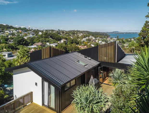 Elevated exterior view of a contemporary dark-clad residence with a sharply pitched corrugated metal roof, timber deck, extensive vertical screening, and rooftop-style planted terraces. The home overlooks a leafy suburban hillside and a broad coastal waterway.