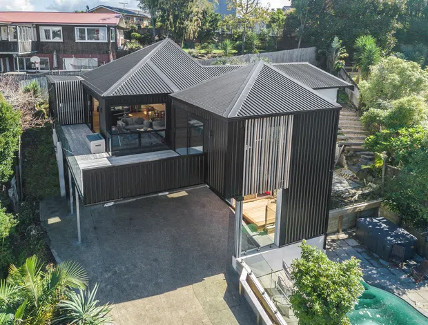Aerial view of a contemporary dark-clad residential building on a sloped, landscaped site. The home has multiple connected pitched roof volumes with corrugated metal roofing, vertical dark cladding, extensive glazing, elevated sections, and a large paved forecourt or terrace. A small pool and outdoor seating area are visible at the lower right, with neighbouring homes and gardens beyond.
