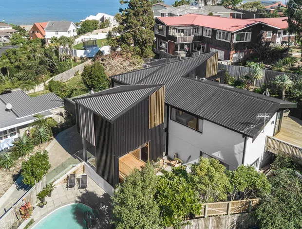 Elevated aerial view of a contemporary multi-level house with dark corrugated metal roofing and cladding, contrasting light plaster walls, timber screening, and expansive glazing. The home sits on a landscaped suburban coastal site with a swimming pool, timber decks, neighbouring houses, and the sea visible in the background.