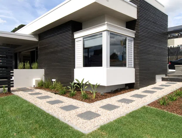 Exterior view of a contemporary multi-level home with crisp white rendered volumes, dark horizontal cladding, projecting flat roof planes, and a prominent corner window. A landscaped front yard includes gravel paths with square stepping stones, garden beds, and lawn.