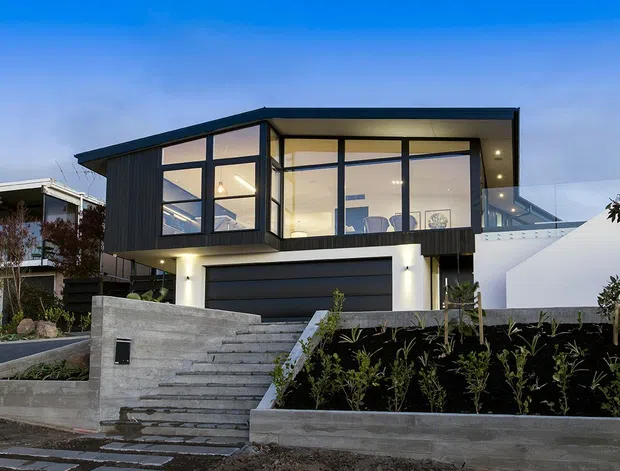 Contemporary two-storey residential house viewed from the street at dusk, featuring a strongly cantilevered upper level, extensive floor-to-ceiling glazing, dark vertical cladding, crisp white rendered walls, a large integrated garage, and a landscaped stepped concrete entry.