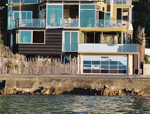 Exterior view of a multi-level waterfront house photographed from the water. The home has extensive turquoise-tinted glazing, projecting balconies with glass balustrades, dark horizontal cladding, a flat projecting roof, and a garage at ground level. Dense vegetation rises behind the property, while a rocky shoreline and water occupy the foreground.