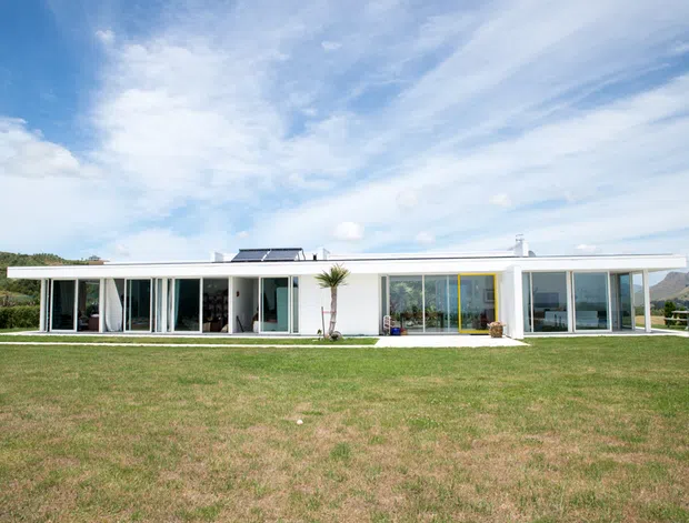 A long, low-slung single-storey modern house set within an expansive lawn, with a predominantly white facade, extensive floor-to-ceiling glazing, slender framing, and a shallow flat roof. The house opens toward a broad landscaped foreground, with rolling hills visible in the background and a bright partly cloudy sky.