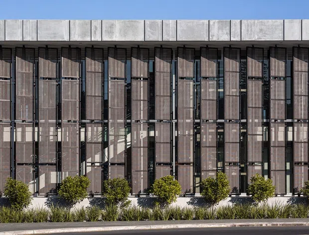 A contemporary multi-storey building facade with a rhythmic grid of tall glazed openings, dark vertical metal framing, and projecting vertical sun-shading fins. A pale concrete parapet or roof edge caps the facade, while a narrow landscaped planting strip with clipped shrubs and grasses lines the base. The image is an exterior architectural elevation photographed in bright daylight.