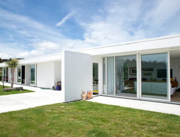 A low-slung, single-storey white modernist house with a flat roof and extensive floor-to-ceiling glazing. The building forms a linear courtyard-facing composition with white rendered walls, a broad concrete-paved terrace or walkway, manicured lawn, and visible open-plan living interiors through the glass. The bright exterior is photographed under a partly cloudy blue sky.
