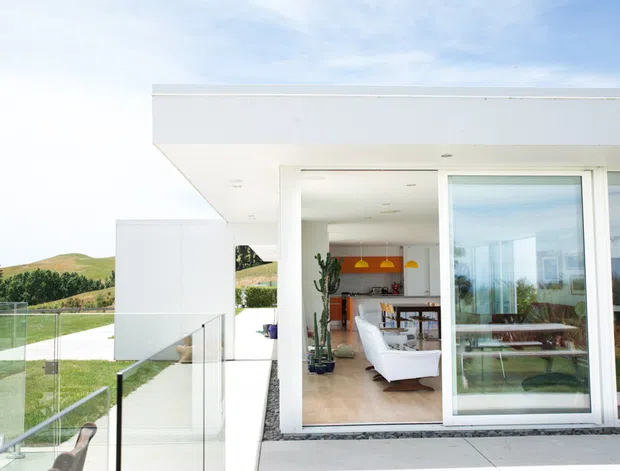 Exterior view of a modern, flat-roofed white residence with expansive floor-to-ceiling glazing and a glass balustrade. Through the open glazing, a bright open-plan living and dining area is visible, with contemporary furnishings, timber flooring, and a colourful kitchen or feature wall in the background. The property overlooks landscaped grounds and rolling hills.