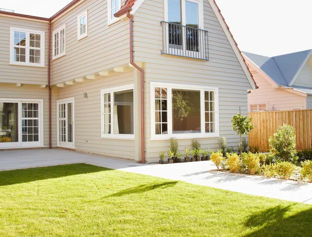 Bright exterior view of a two-storey suburban house with cream horizontal weatherboard cladding, white-trimmed multi-pane windows, a steep front-facing gable roof with red tile roofing, and a small black metal balcony. A paved terrace and neatly landscaped lawn with low shrubs occupy the foreground, with neighbouring pitched-roof homes visible in the background.