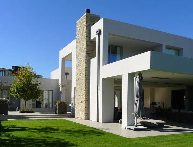 Exterior view of a large modern two-storey residence with crisp white rendered walls, deep rectangular overhangs, recessed balconies, and extensive open glazing. A tall stacked-stone chimney or feature wall forms a strong vertical element at the front. The house opens onto a paved terrace and manicured lawn, with a covered outdoor entertaining area and poolside-style lounge furniture visible beneath the upper volume.