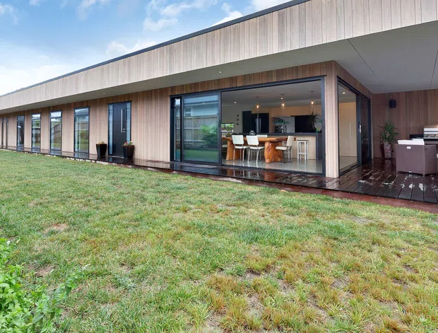 Wide exterior view of a long, single-storey contemporary home with a low-pitched roof, extensive floor-to-ceiling glazing, vertical timber cladding, and a covered timber deck. The open glazing reveals a kitchen and dining area, while an outdoor seating area is visible at the right.