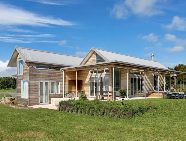 A single-storey contemporary rural residence viewed from the garden, with weathered timber-clad walls, pale corrugated pitched roofs, extensive glazed doors and windows, and a covered outdoor terrace. The house sits in a broad landscaped lawn with mature trees beyond, under a bright blue sky.