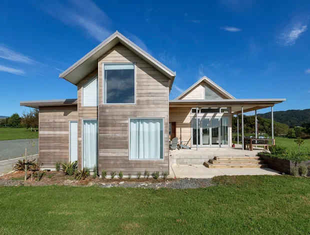 Exterior photograph of a contemporary timber-clad house set in a landscaped rural or semi-rural setting. The building has prominent gabled roof forms, extensive glazing, a covered patio or veranda, broad steps, and a mix of horizontal weatherboard cladding and light masonry paving.