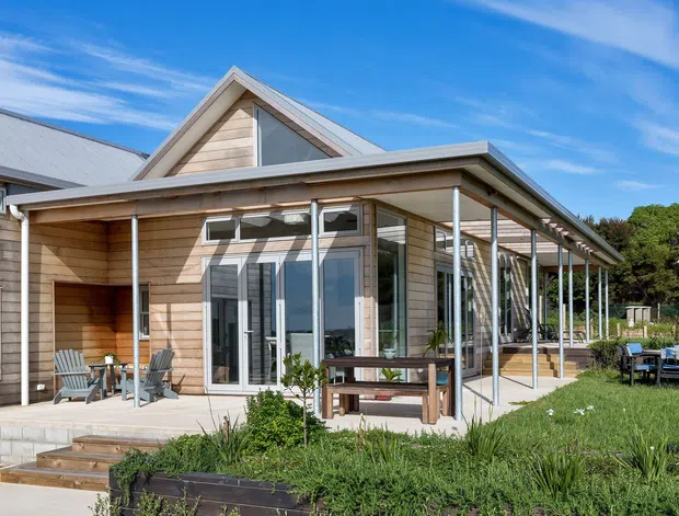 Exterior view of a contemporary single-storey timber-clad house with prominent gabled roof forms, extensive glazing, and a long covered outdoor veranda supported by slender posts. A paved terrace, outdoor seating, timber steps, lawn, and garden planting surround the building.