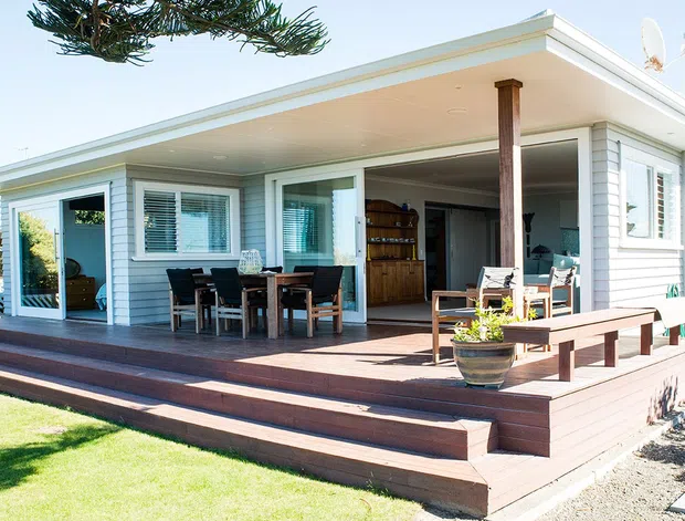 Single-storey coastal-style house with a broad covered outdoor deck, wide sliding glass doors, white horizontal weatherboard cladding, and a low-pitched flat roof. The deck includes outdoor dining and lounge furniture, planter pots, and broad timber steps leading to a lawn.