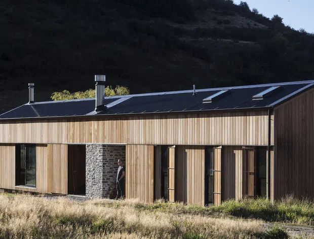 A compact single-storey timber-clad house set against a steep, wooded hillside. The facade features vertical natural timber boards, dark-framed windows and doors, a projecting stone-clad chimney or entry element, and a simple pitched metal roof with skylights and chimneys. Dry grass and native vegetation surround the building, giving it a secluded rural or bush setting.