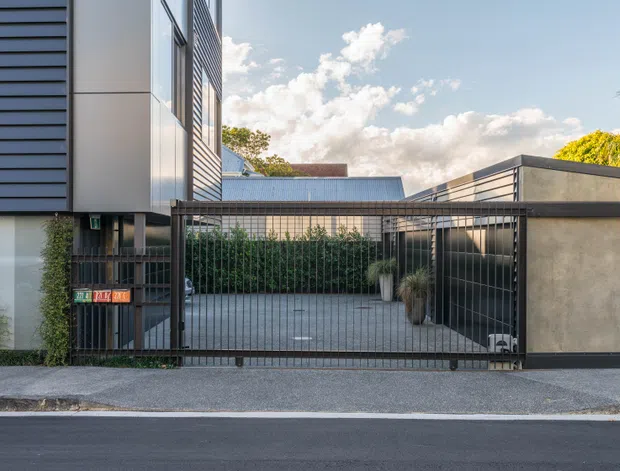 Street-level view of a contemporary residential property frontage with dark horizontal cladding, rendered wall panels, metal security fencing and sliding vehicle gate. A paved driveway leads toward a low, flat-roofed structure, with planted screening along the boundary and a neighbouring modern building visible behind.
