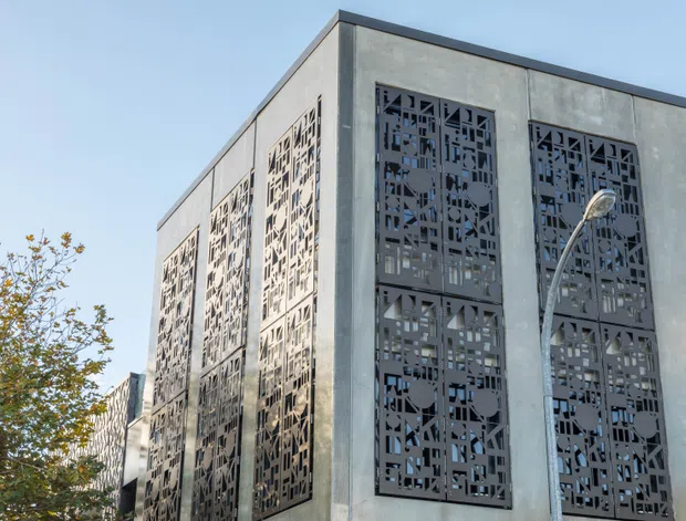 Exterior view of a contemporary multi-storey building with a stark rectangular concrete facade. Tall, narrow decorative perforated metal screens cover the windows, creating a geometric patterned elevation. A curved streetlight and some trees appear in the foreground under a clear sky.