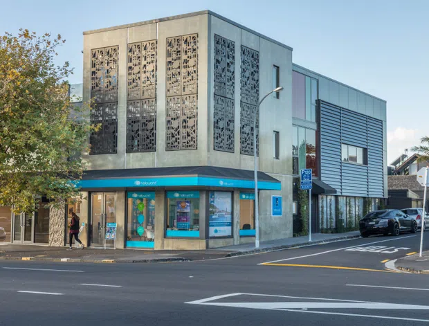 Street-level view of a contemporary mixed-use commercial building on a city corner. The ground floor has a bright turquoise retail or service tenancy, while the upper facade features decorative perforated metal screening and the adjoining section uses dark horizontal cladding with large windows. Urban streets, signage, mature trees and neighbouring buildings surround the site.