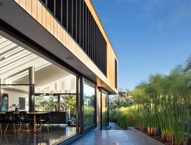 Contemporary two-level residential extension with extensive floor-to-ceiling glazing opening onto a landscaped outdoor area. The exterior features warm vertical timber cladding, dark vertical screening, and a broad roof overhang. Inside, a bright open-plan dining and kitchen area is visible, with polished dark flooring, pendant lights, a dining table, and modern furnishings. The image is taken from an outdoor terrace or walkway looking toward the glazed facade and garden.