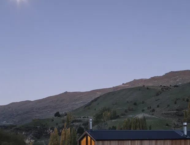 A compact single-storey timber-clad house sits in a rural valley beneath rolling, sparsely vegetated hills. The dark pitched roof, warm vertical timber walls, large glazed gable end and illuminated interior create a strong contemporary cabin-like form. Tall trees and a low red fence line surround the property, with a clear blue dusk sky above.