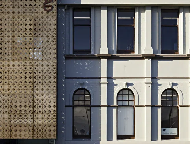 Street-level view of a narrow historic-style building facade with ornate patterned tile cladding on the left and a pale rendered masonry facade on the right. The facade features strong vertical pilasters, cornice-like horizontal bands, rectangular upper windows, and arched lower windows with dark frames. A small sign is visible in one lower window.