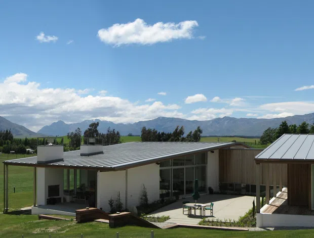 Wide exterior view of a low-slung contemporary house set in an open rural landscape, with a flat-roofed central pavilion, a separate pitched metal-roofed wing, extensive glazing, timber-clad walls, and an outdoor paved terrace. Mountains, fields, trees, and a bright blue sky form the background.