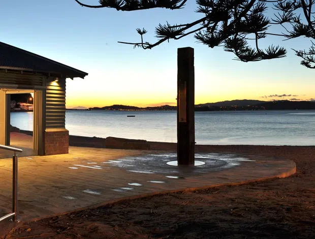 A dusk exterior view of a small timber-clad waterfront pavilion or beach facility beside a sandy shoreline. The building has a low dark hipped roof, warm horizontal timber walls, illuminated windows, and an accessible metal-railed pathway leading toward the water. A pier extends across the bay, with trees framing the scene.