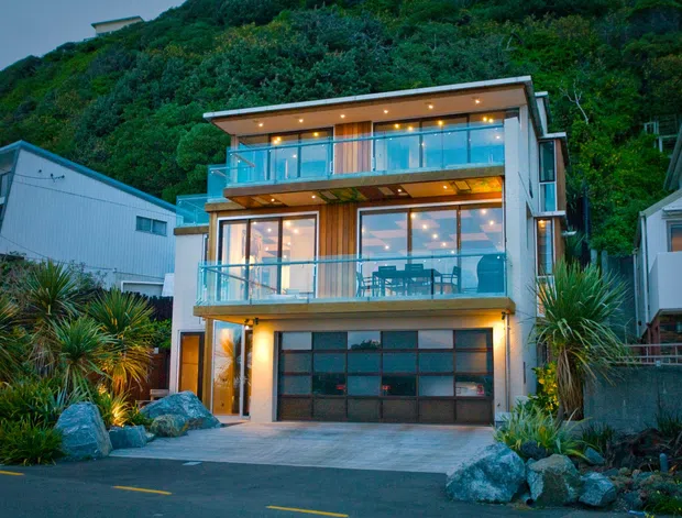 A modern multi-storey hillside house photographed at dusk, with extensive floor-to-ceiling glazing, illuminated interiors, timber-toned cladding, glass-balustraded balconies, and a ground-level garage. The home is set into a densely vegetated steep site with landscaped native-style planting and rocks, alongside neighbouring houses.