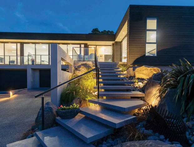 Twilight exterior view of a contemporary multi-level home on a landscaped sloping site. The house features dark horizontal cladding, extensive glazed living areas, a projecting upper volume, an attached garage, and a broad exterior stairway with modern metal balustrades. Rock landscaping and architectural lighting frame the entry.