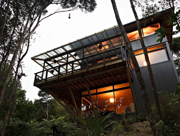 Exterior view of a multi-level elevated dwelling set among dense native bush. The building features a projecting timber-framed deck, extensive glazing, corrugated dark metal cladding, and a translucent or glazed roofed terrace structure. Warm interior lighting glows through the windows, contrasting with the surrounding forest.