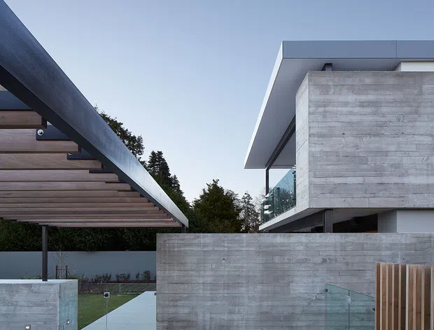Exterior architectural view of a contemporary residence with strong horizontal geometry, exposed board-formed concrete walls, broad flat roof overhangs, a timber-slatted carport or covered outdoor structure, glass balustrades, and a timber deck. The composition emphasizes the building facade and built landscape elements against a clear sky and trees.