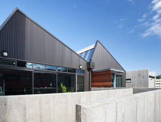 Contemporary commercial or residential building exterior with a strong repeated gable roof form, dark vertical ribbed cladding, extensive glazed openings, exposed concrete retaining walls, and a warm timber-clad recessed entrance. The image is photographed outdoors under a clear sky, with a clean modern architectural composition.