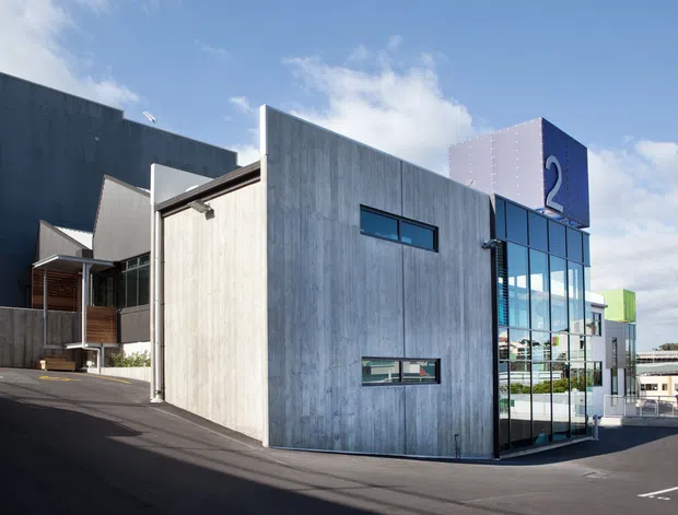 Exterior view of a contemporary multi-level commercial building on a sloped paved site. The facade combines large weathered grey concrete panels, extensive blue-tinted glazing, dark wall sections, and colourful geometric rooftop or facade elements. A covered entry and projecting architectural volumes are visible, with adjacent commercial buildings in the background.