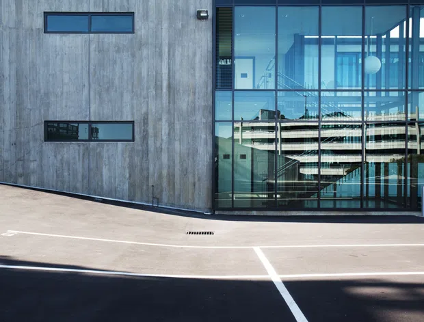 Exterior view of a contemporary multi-storey building with a tall glazed stairwell or circulation core beside a textured grey concrete facade. Narrow horizontal windows punctuate the solid wall, while the reflective glazing reveals internal stairs and surrounding urban buildings. The foreground is a paved driveway or parking area with painted white lines.
