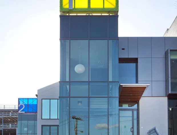 Exterior photograph of a contemporary multi-storey commercial or institutional building. The facade combines extensive blue-tinted floor-to-ceiling glazing, dark metal framing, white and grey panel cladding, and a prominent rooftop structure with vivid lime-green and yellow translucent panels. A glazed entrance is accessed by broad stairs, with chain-link fencing in the foreground and another numbered glazed volume visible behind.