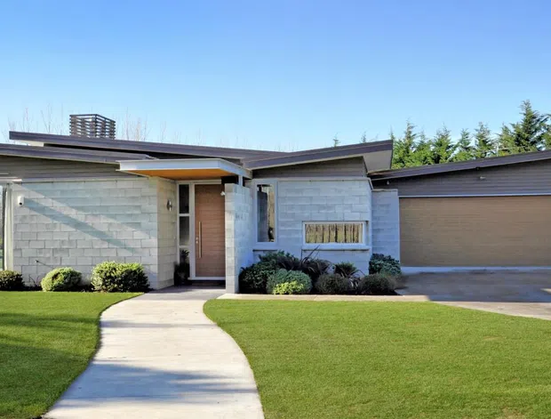 Single-storey contemporary residence viewed from the front, with a low-slung flat roof, light masonry walls, timber-toned entry door, broad glazing, and an attached double garage. A curved concrete path leads through a manicured lawn to the recessed entry, with clipped planting and screening trees around the property.