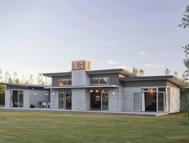Single-storey contemporary residence viewed from the landscaped lawn, with a low horizontal form, flat roof planes, extensive floor-to-ceiling glazing, light grey masonry walls, dark trim, and a central chimney or feature volume. Mature trees and a broad lawn frame the exterior.
