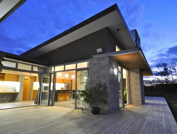 Twilight exterior view of a contemporary single-storey home opening onto a broad timber deck. The house features strong horizontal roof planes, extensive floor-to-ceiling glazing, dark horizontal cladding, pale masonry walls, and warm interior lighting visible through the glass. A kitchen and dining area are visible inside, while the deck and landscaped perimeter frame the exterior.