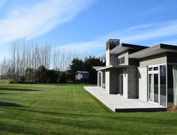 A low, modern single-storey house set beside a broad, neatly mown lawn. The building has a flat, layered roofline, pale grey masonry or concrete-block walls, dark horizontal cladding accents, extensive glazed doors and windows, and a paved terrace along the facade. Leafless trees and open grounds surround the property.