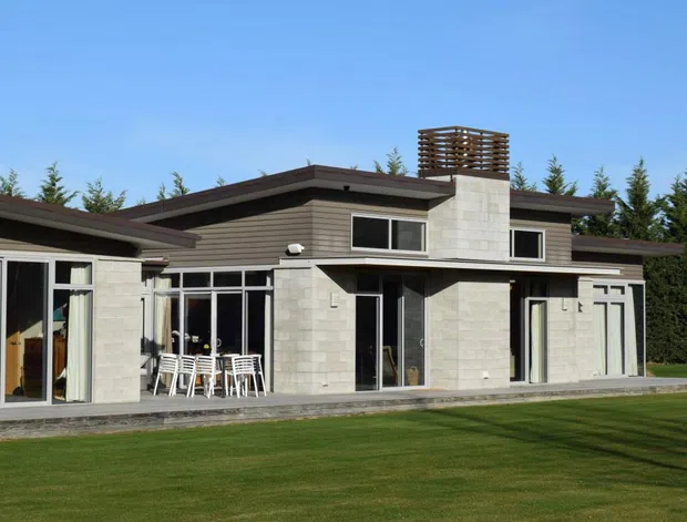 Single-storey contemporary residence viewed from the rear, with a low-pitched roof, pale masonry walls, extensive floor-to-ceiling glazing, multiple patio doors, and a broad lawn. A tall central chimney or exhaust feature is finished with a distinctive open timber-like screen. Outdoor dining furniture is visible beneath the covered terrace.