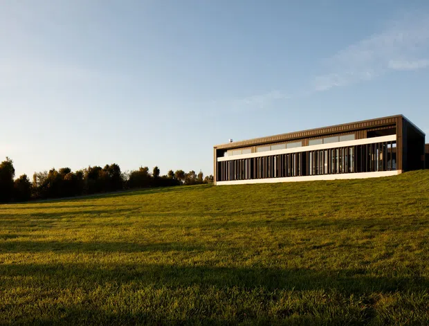 A long, low modern rectangular building sits prominently on a gently sloping grassy field. The facade features extensive floor-to-ceiling glazing, strong horizontal bands, dark vertical screening or fins, and a flat or very low-pitched roof with a projecting upper edge. The scene is photographed in warm late-day light with open lawn and a line of trees in the background.