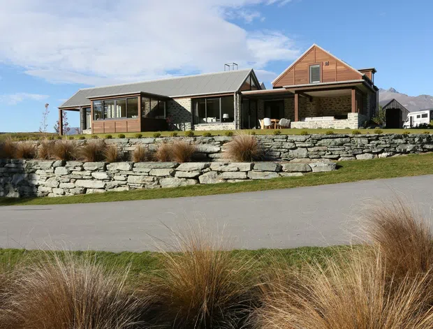 A single-storey contemporary residential home set on a landscaped, gently sloping site. The house combines dark timber cladding, grey metal roofing, extensive glazing, and a prominent timber-clad gabled volume, with broad stone retaining walls and native tussock-style planting in the foreground. Mountain terrain is visible in the background under a bright sky.